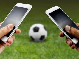 Two people hold phones while there is a football on the grass in front of them