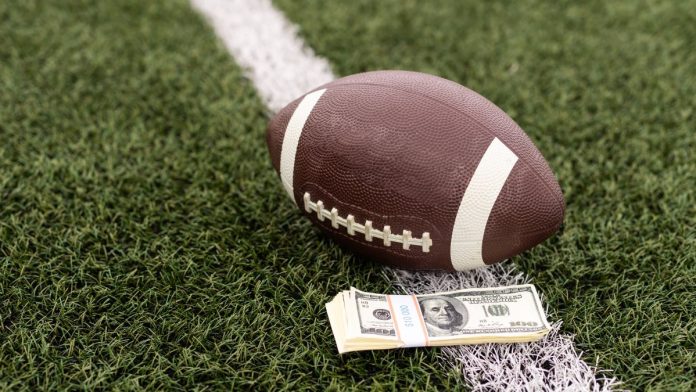 Kalshi-took-$441M-in-trading-volume-in-NFL-Week-1,-says-CEO An NFL football lying on a football field next to a stack of U.S. banknotes