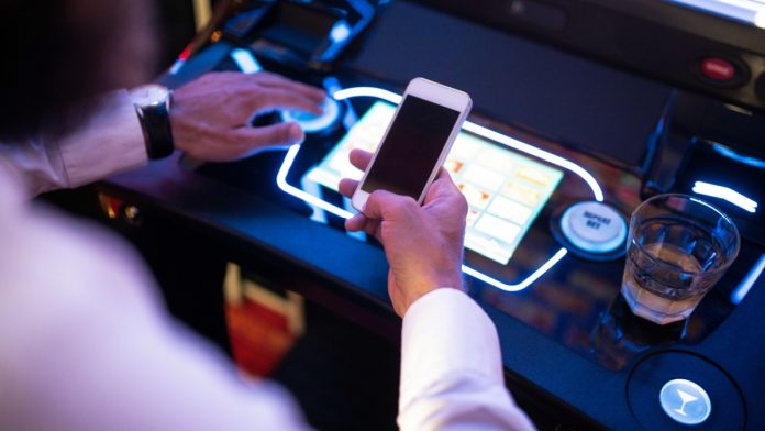 Aristocrat-Acquires-Awager-Live-Slot-Streaming Man has phone at slot machine as Aristocrat acquires live slot streaming company Awager.