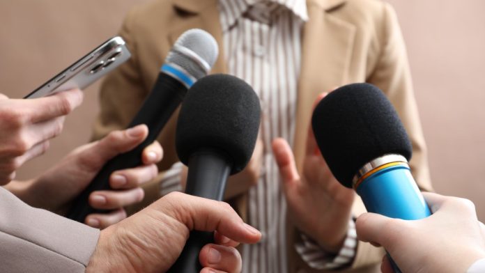 Several people holding up microphones to a person as if questioning them