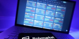 The Polymarket logo on a phone with trading markets on a laptop screen in the background