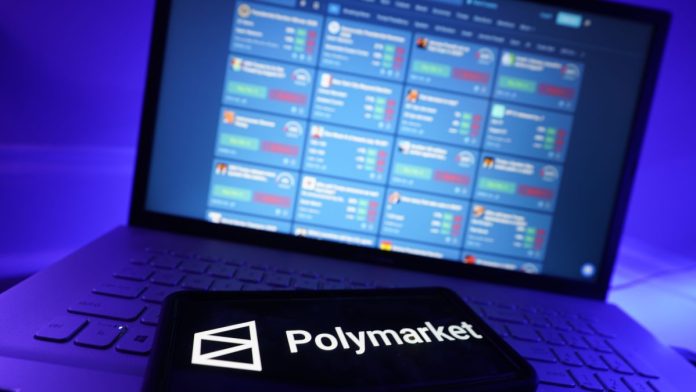 PrizePicks'-Polymarket-partnership-will-vary-from-state-to-state The Polymarket logo on a phone with trading markets on a laptop screen in the background